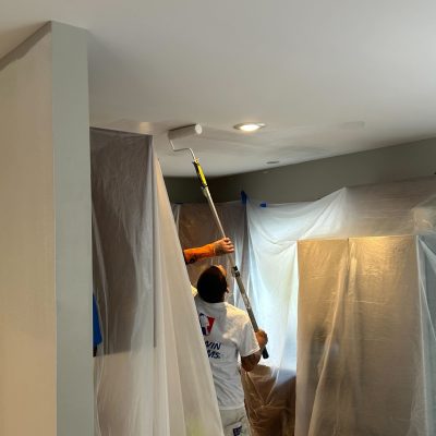 A house painter, wearing a white painter's suit and a blue baseball cap, paints the exterior of a Victorian house.