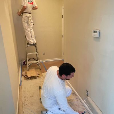 Two painters working together in a hallway, one on a ladder and one rolling near the baseboard.
